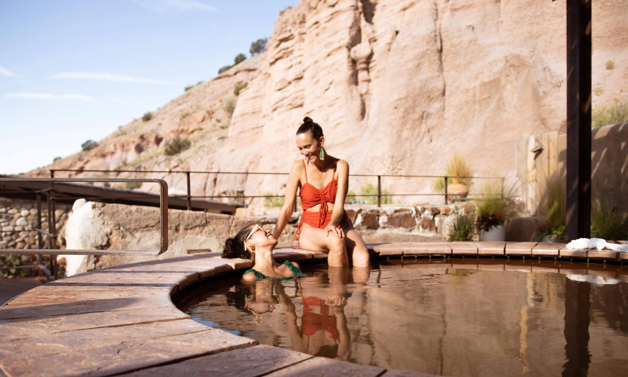 Two women in swimsuits soaking in a round pool with tan cliffs in the background